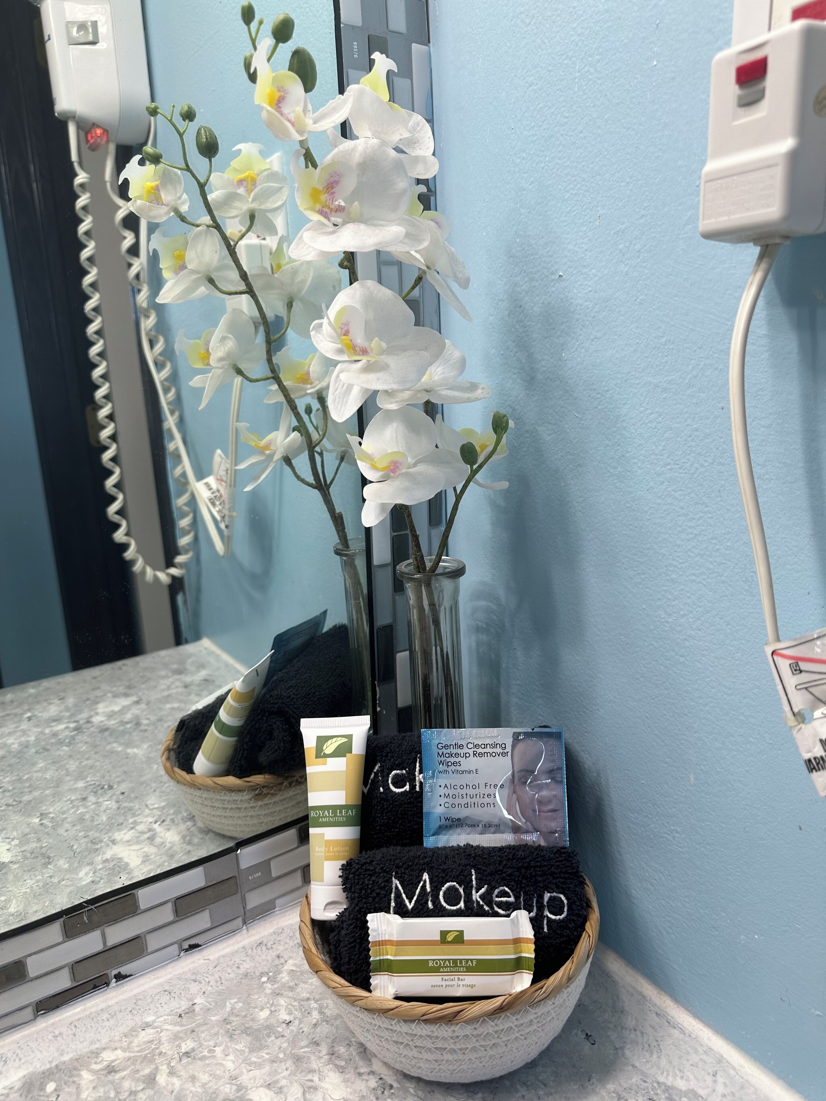 A bathroom vanity with a mirror, a white orchid plant, and a basket containing a facial bar, a tube of body lotion, and a pack of makeup wipes.