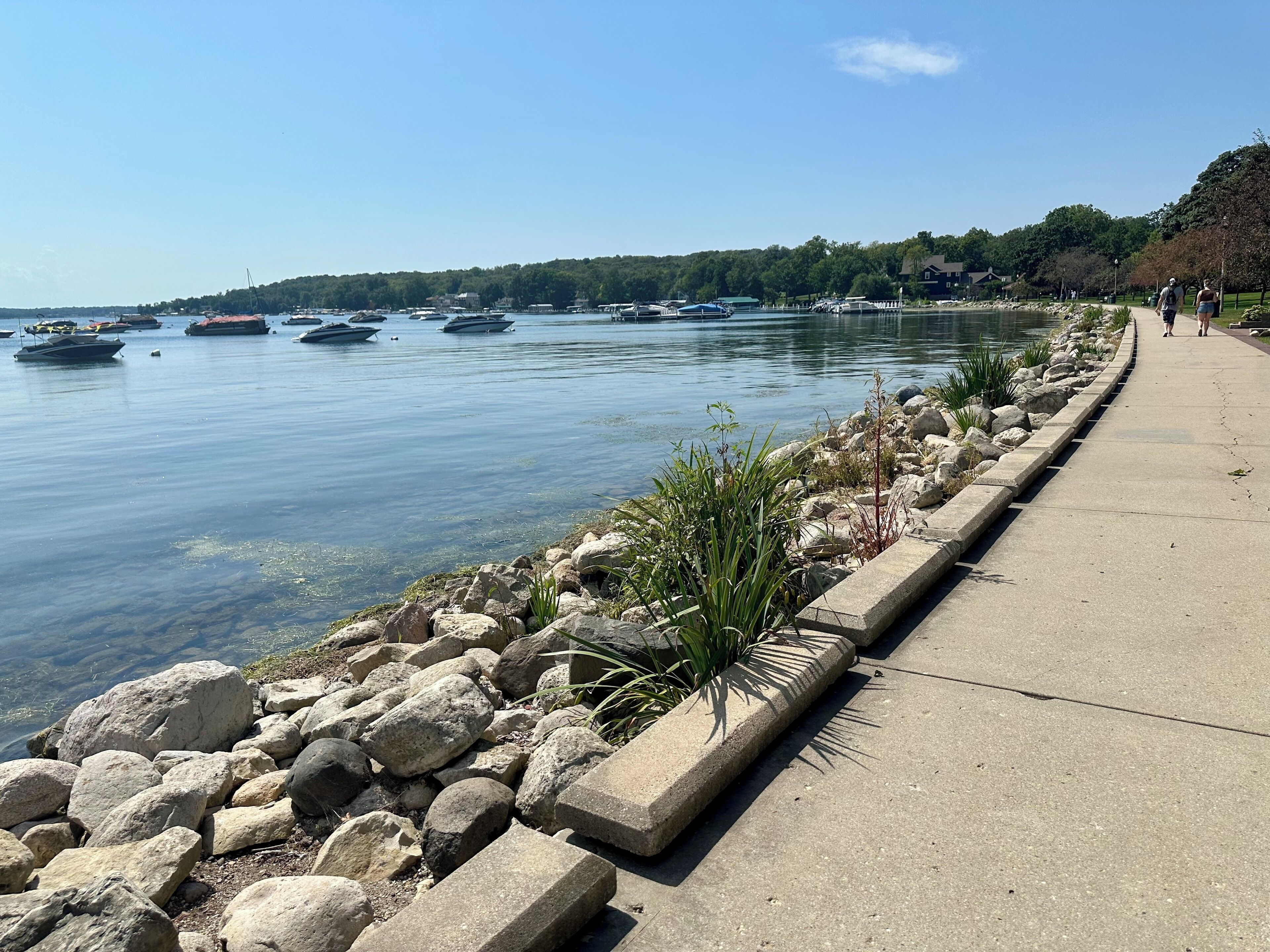 A waterfront promenade with a concrete walkway, rocky shoreline, and boats on the water.