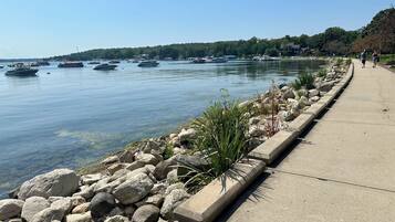 A waterfront promenade with a concrete walkway, rocky shoreline, and boats on the water.