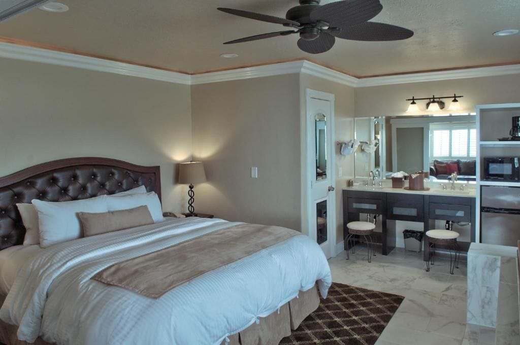 Una camera d'albergo con un letto grande, una ventola a soffitto, un bagno con lavabo e specchio, e un angolo cottura.