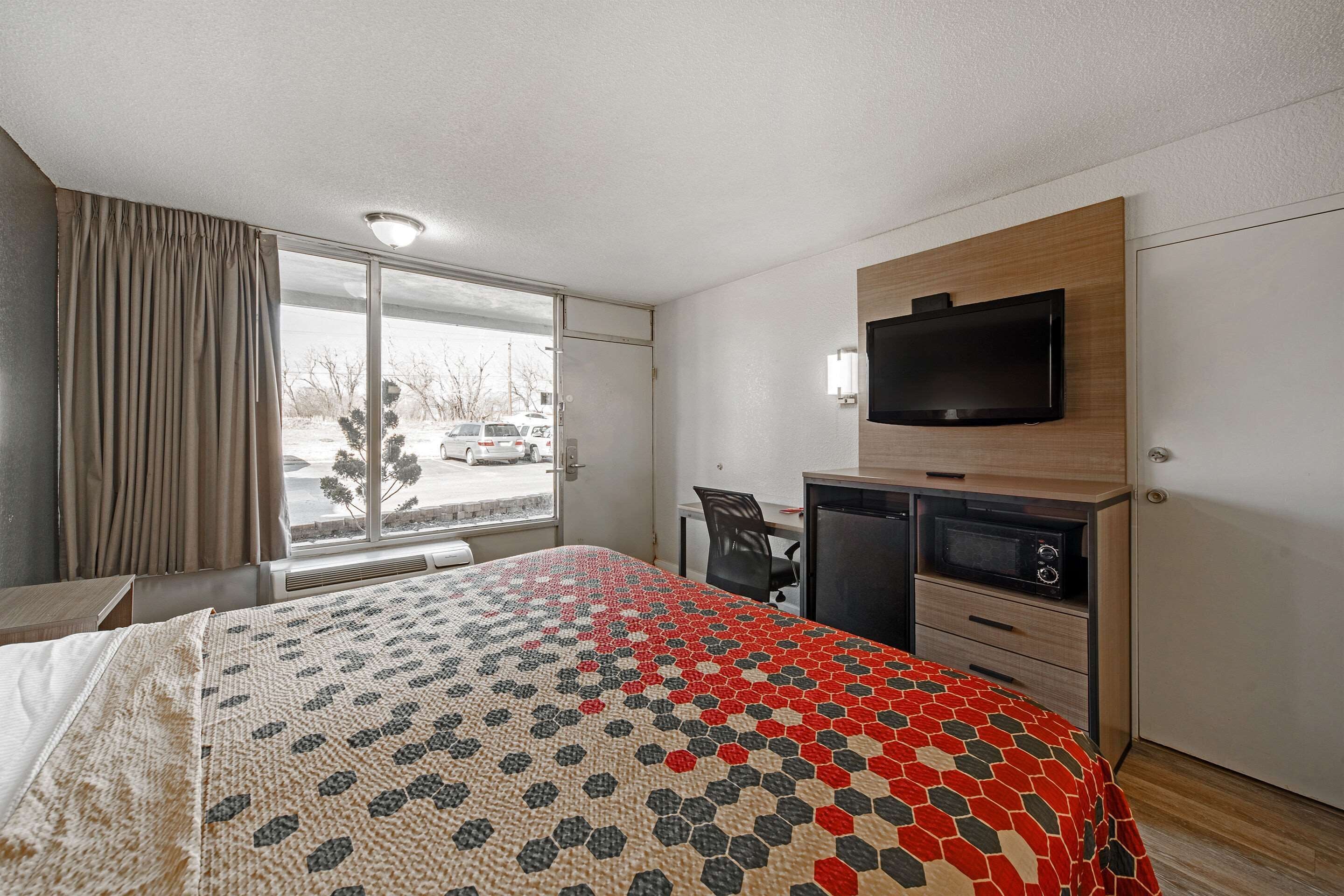A hotel room with a large bed, a desk with a computer, a TV mounted on the wall, and a window with a view of parked cars outside.