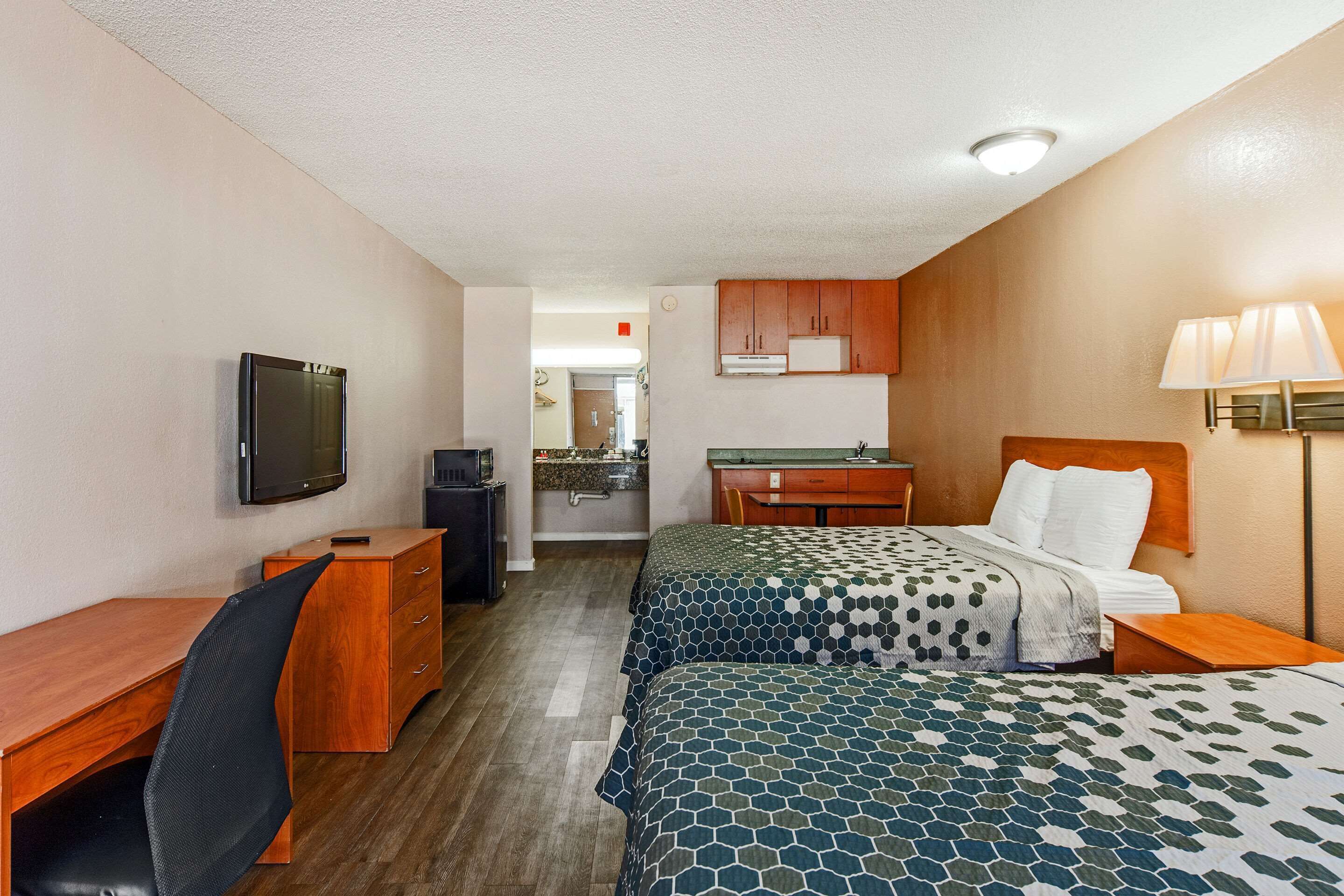 Una habitación de hotel con cama, escritorio, silla, televisor, lámpara y kitchenette.
