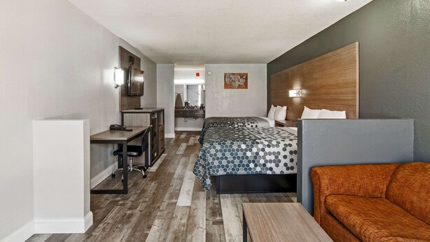 Una habitación de hotel moderna con piso de madera, una cama con colcha estampada, un escritorio con una silla, un sofá y un televisor montado en la pared.
