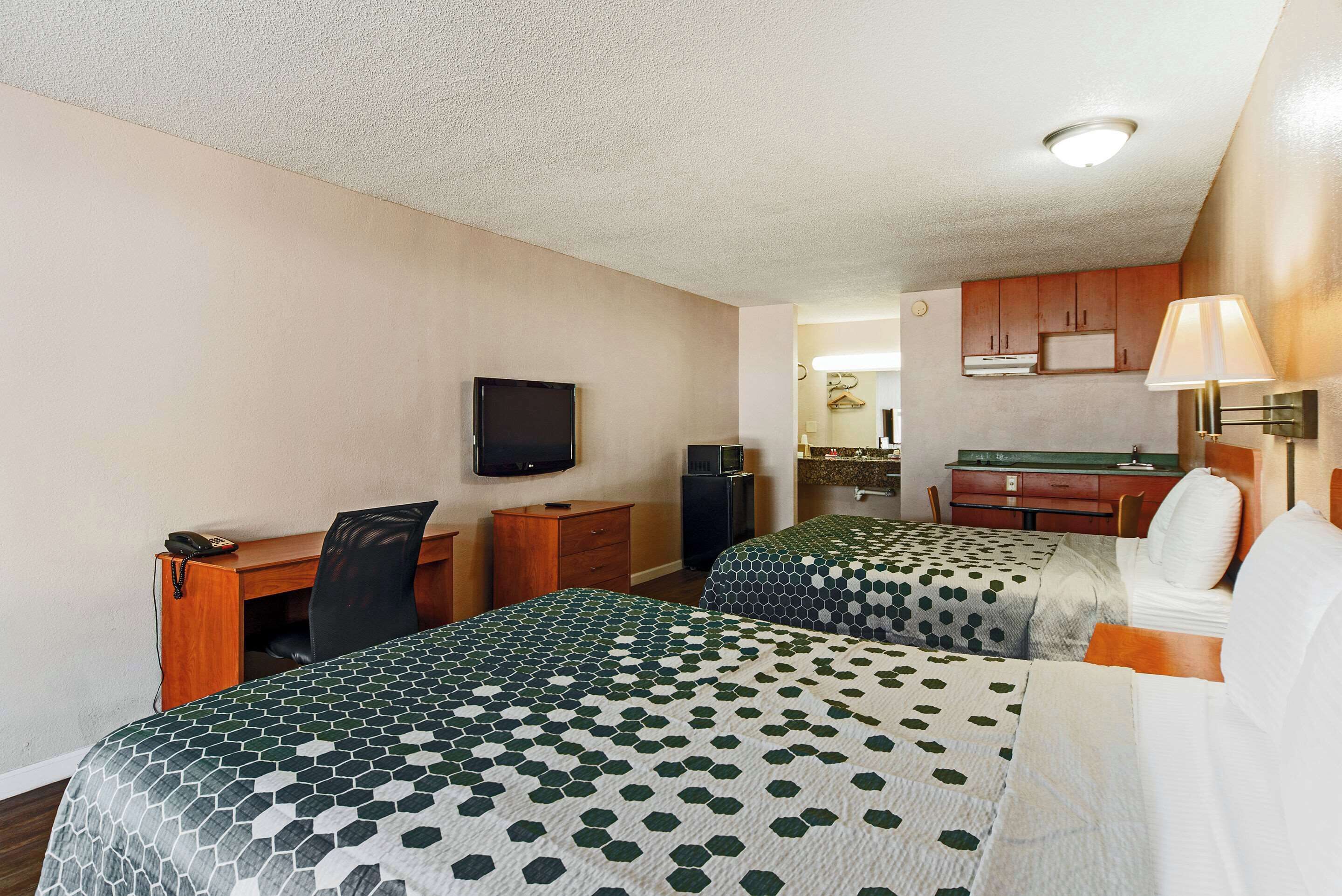 A hotel room with two beds, a desk, a chair, a TV, and a kitchenette.