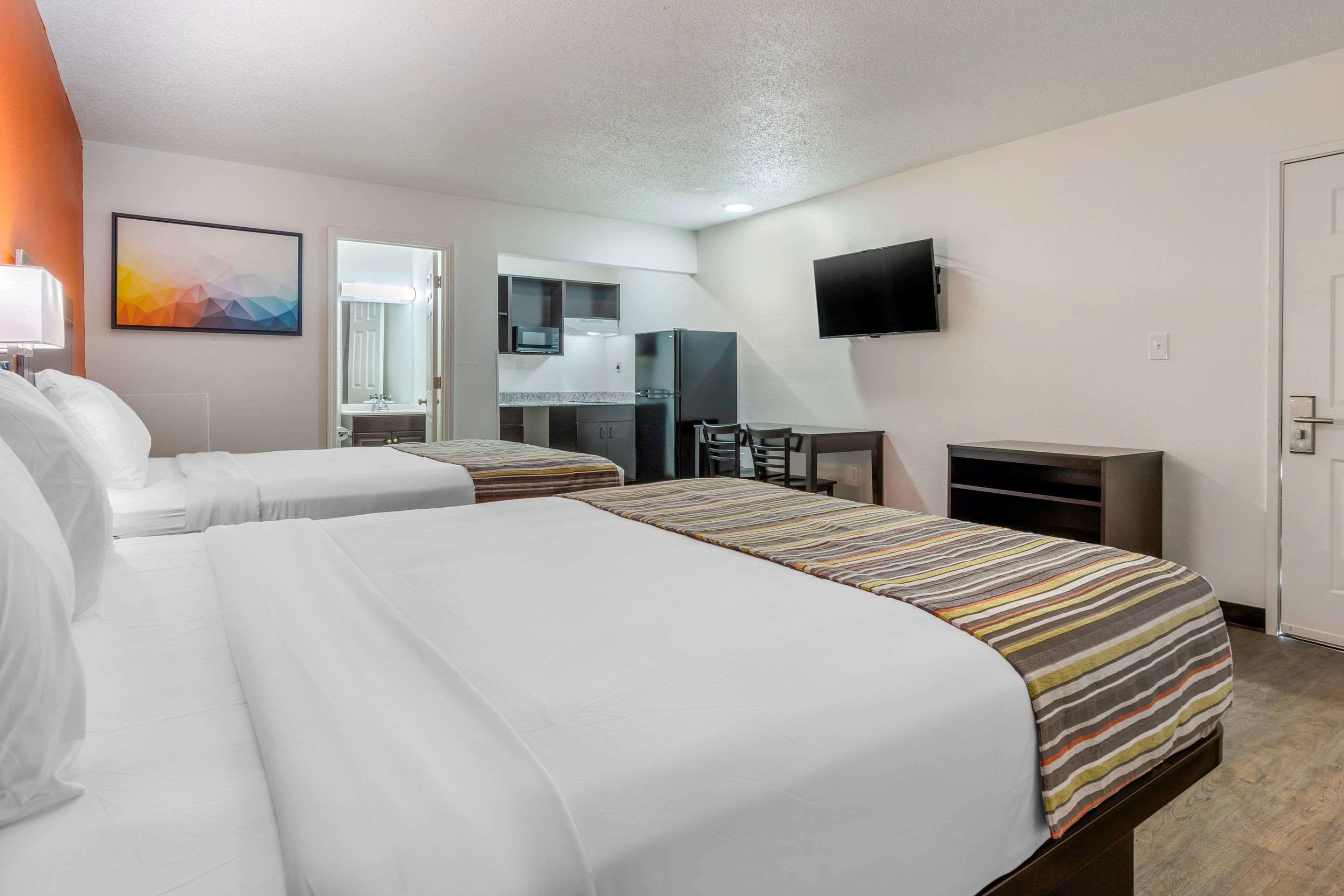 A hotel room with two beds, a kitchenette, a TV, and a bathroom visible through an open door.