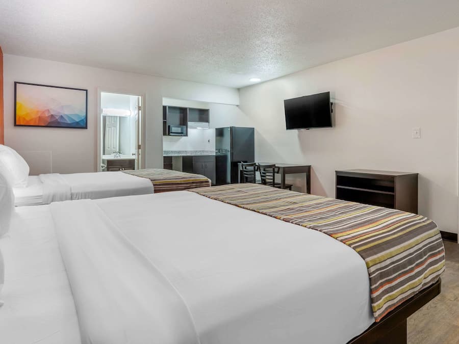 A hotel room with two beds, a kitchenette, a TV, and a bathroom visible through an open door.