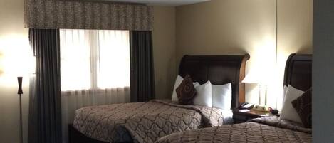 A hotel room with two beds, a large window with curtains, a standing lamp, and a bedside table with a lamp.
