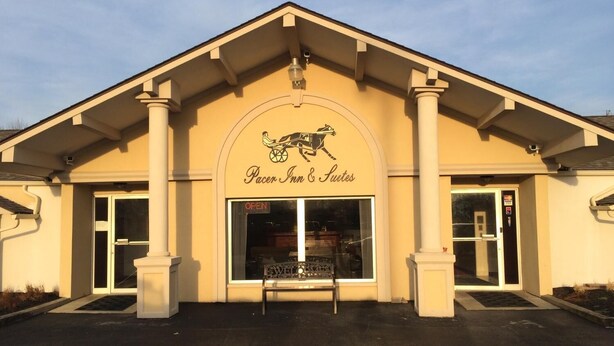 Das Außere eines Hotels namens "Pacer Inn & Suites" mit klassischer Architektur, mit einem zentralen Eingang, einer Bank, großen Fenstern und einem Schild, das anzeigt, dass die Einrichtung geöffnet ist.