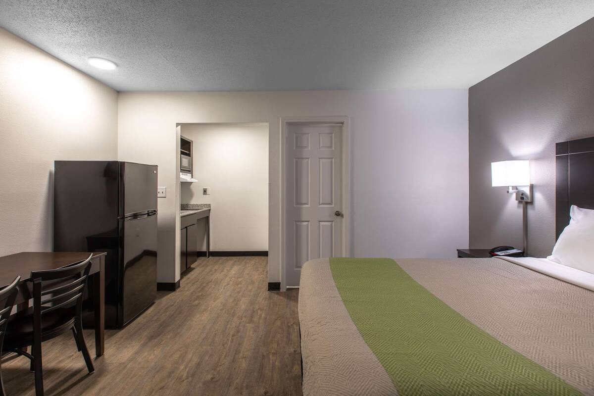 A hotel room with a bed, a desk, a refrigerator, a chair, and a nightstand with a lamp.