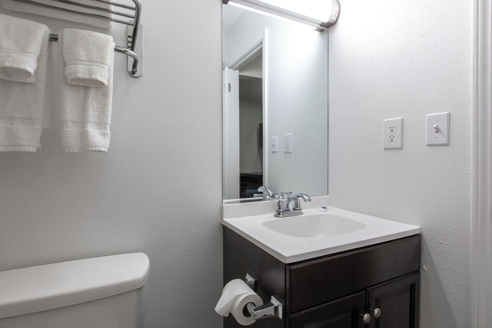 Un baño con un lavamanos blanco, un mueble de almacenamiento oscuro y un inodoro. Hay toallas colgadas en un perchero y un espejo sobre el lavamanos.