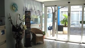 Un área de recepción con una silla de mimbre, un jarrón alto con flores, un reloj y una puerta de cristal que da al exterior.