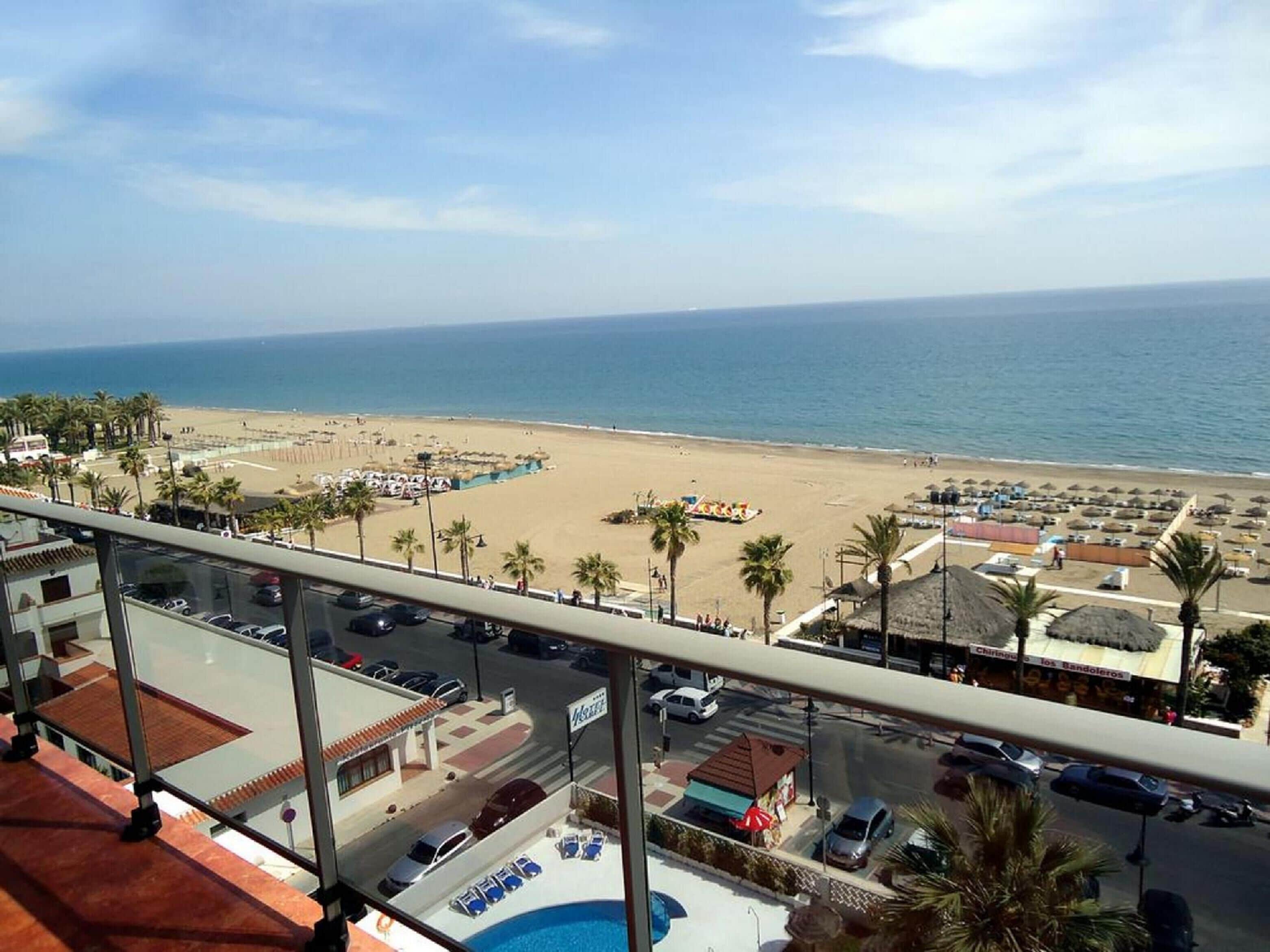 Une vue sur la plage depuis un balcon, avec une piscine, des palmiers et des parasols de plage.