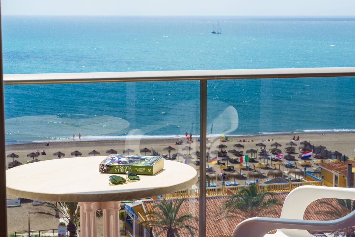A balcony with a view of the beach, a table with books, and a chair.