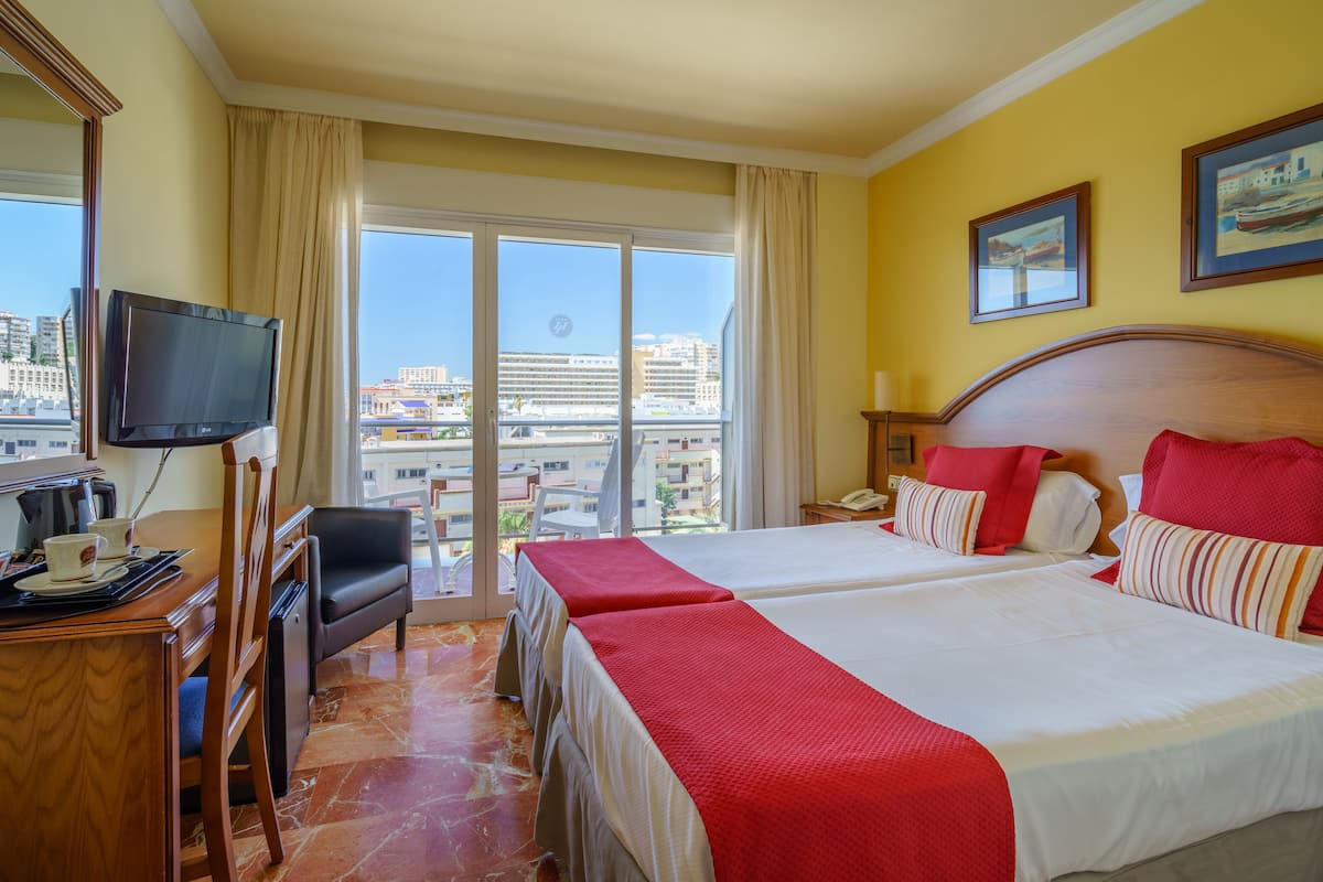 A hotel room with two beds, a TV, a desk with a chair, and a view of the city through large windows.