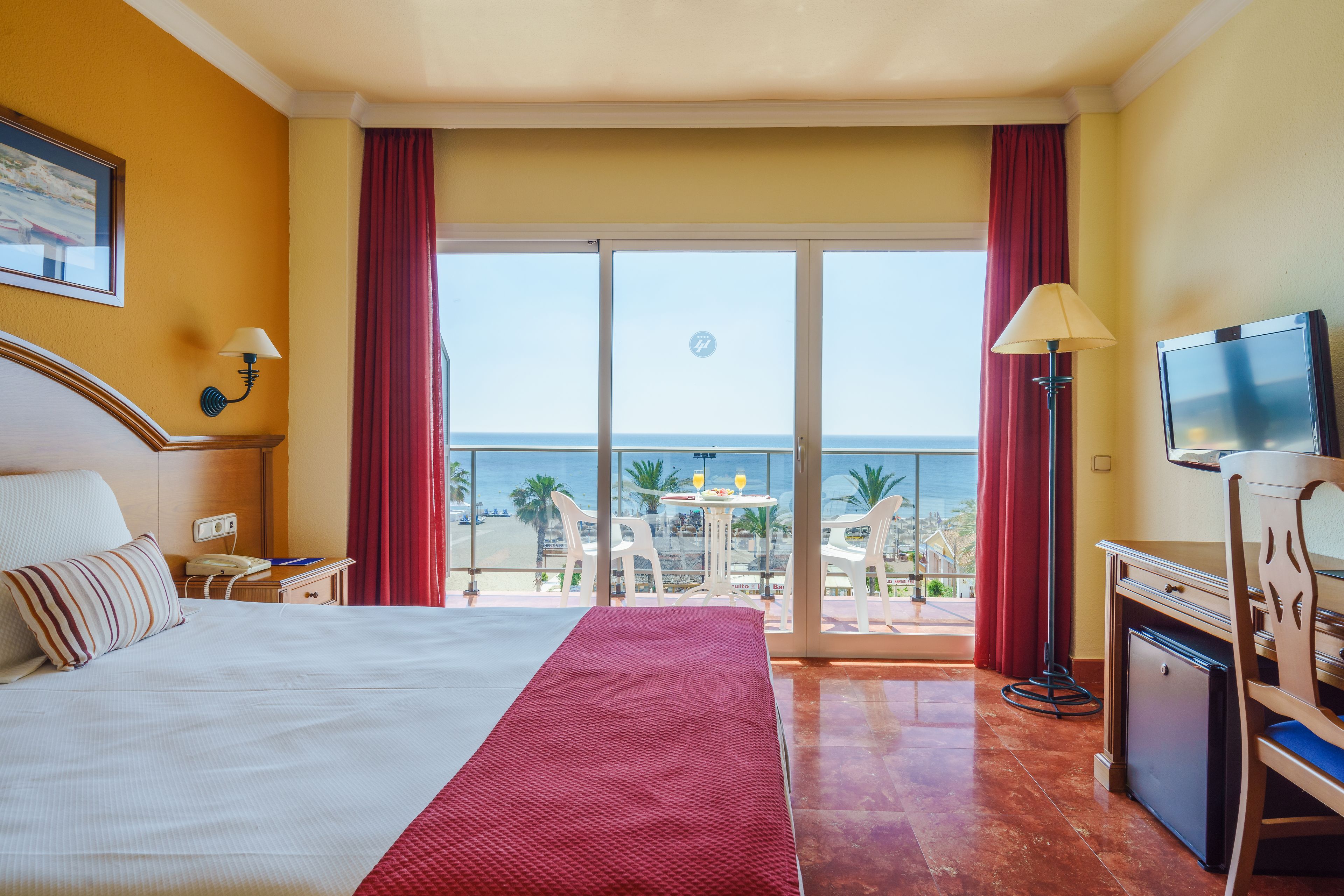 A hotel room with a bed, a desk with a television, a small fridge, and a balcony with a view of the ocean.