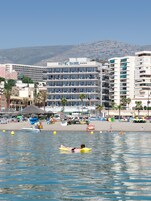 Een hotel met meerdere verdiepingen en balkons, een strand met parasols en ligstoelen, en een waterpartij met mensen en boten.