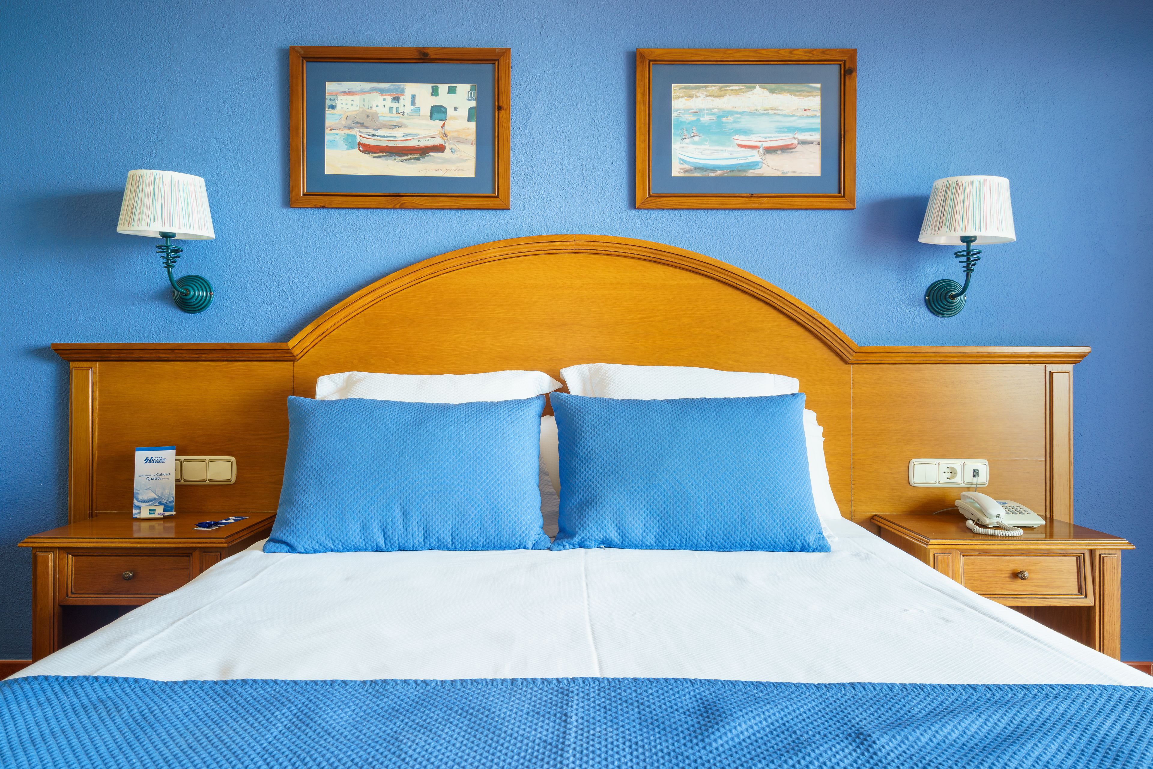 A hotel room with a wooden headboard, two blue pillows, a white bedspread, and two wall-mounted lamps.