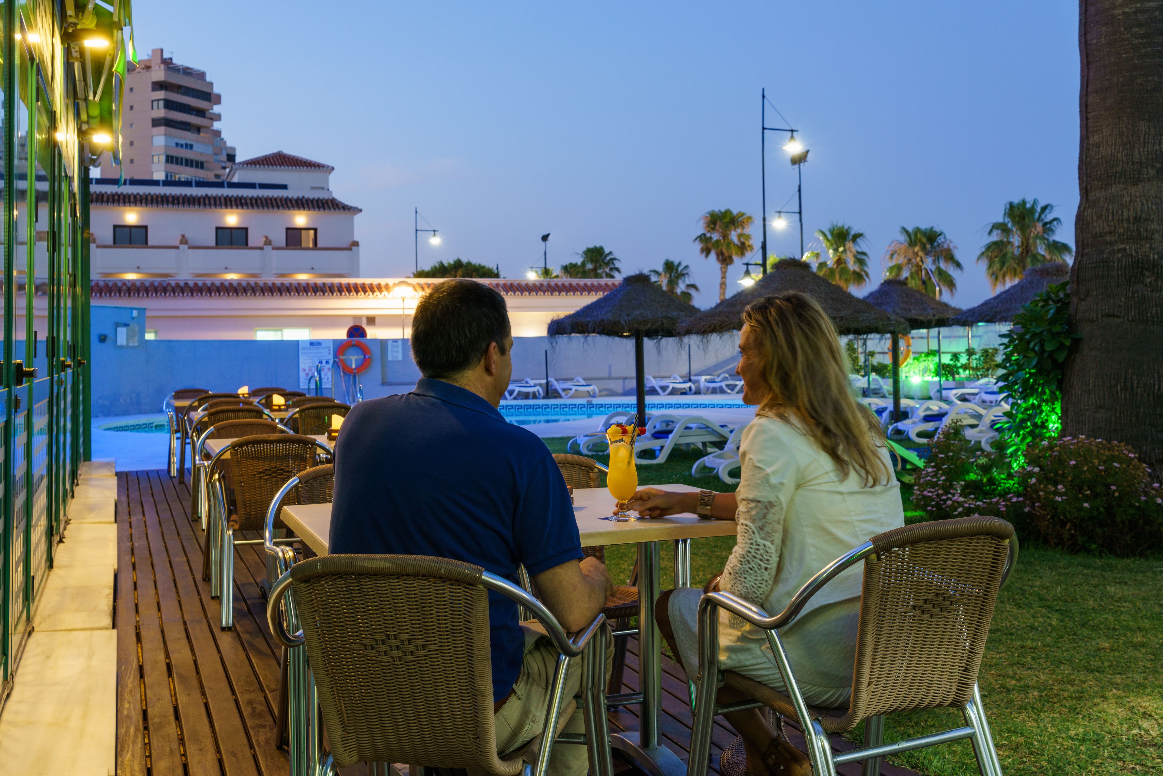 Una pareja cenando en un restaurante al aire libre con vista a una piscina y un estacionamiento.