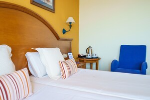 A hotel room with a bed, pillows, a blue armchair, a nightstand with a lamp, and a coffee maker.