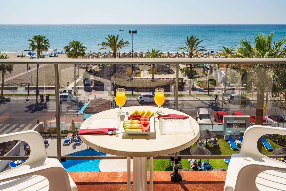 A balcony with a table set for breakfast, overlooking a beach and pool area.