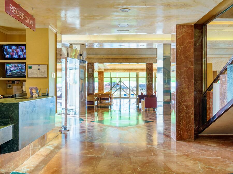 Uma área de recepção de hotel com piso de mármore, balcão de atendimento, telas de televisão e uma escadaria.