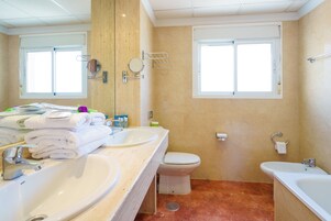Een badkamer met twee wastafels, een toilet en een bad. Handdoeken liggen op de linker wastafel en er is een raam boven het toilet.