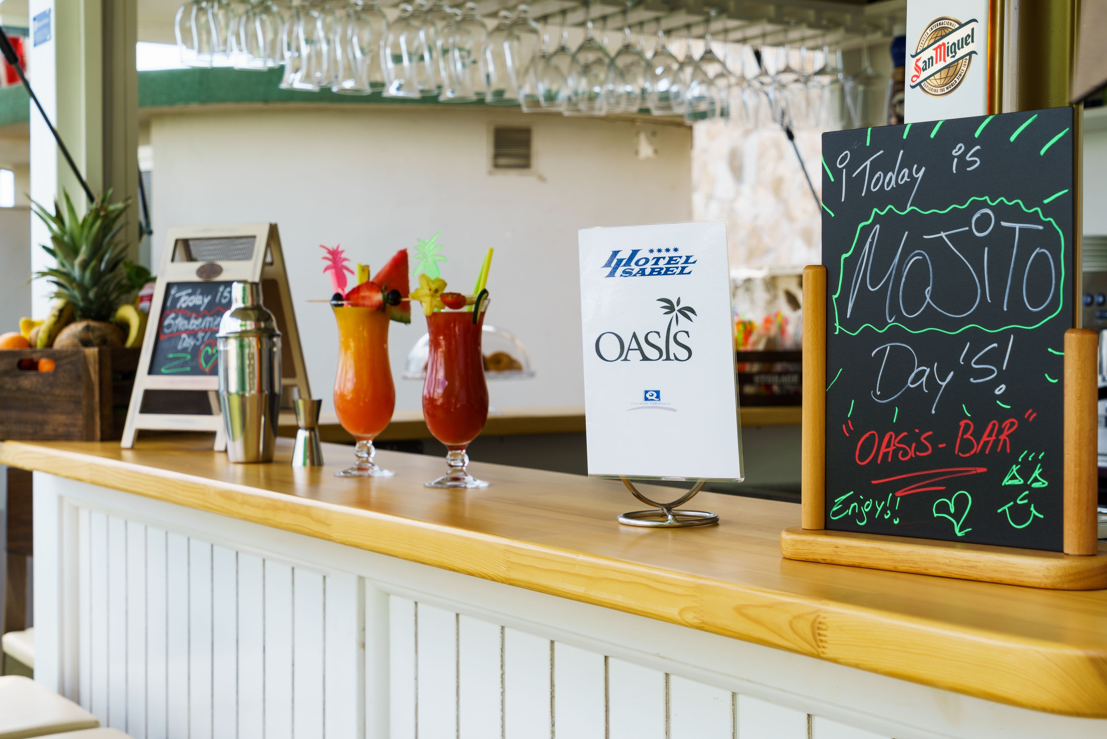 A bar area with a chalkboard sign announcing 'Today is Mojito Day' at Oasis Bar, featuring two cocktails and a sign for Hotel Oasis.