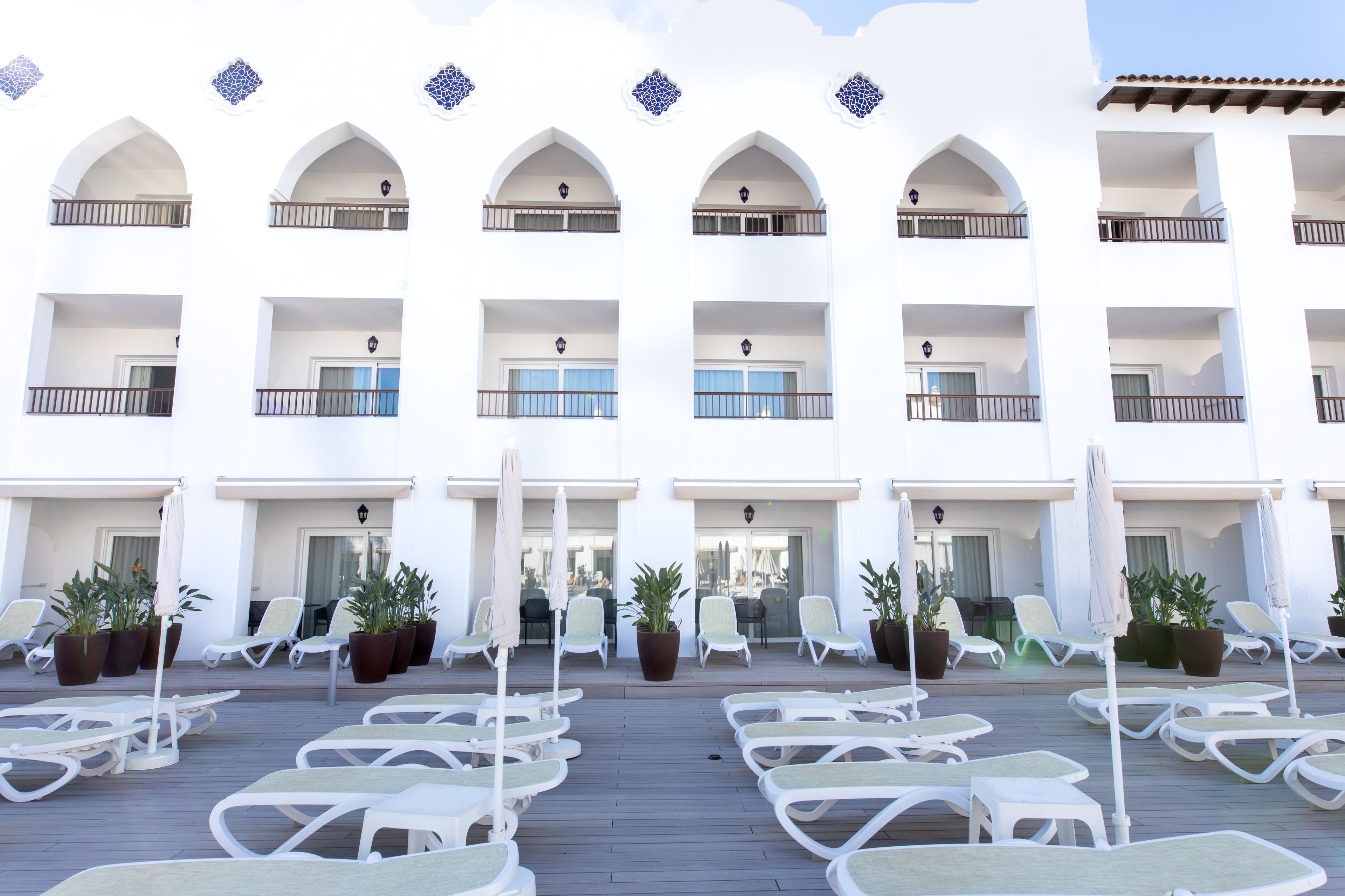 A hotel with multiple balconies, outdoor seating areas with chairs and umbrellas, and potted plants.