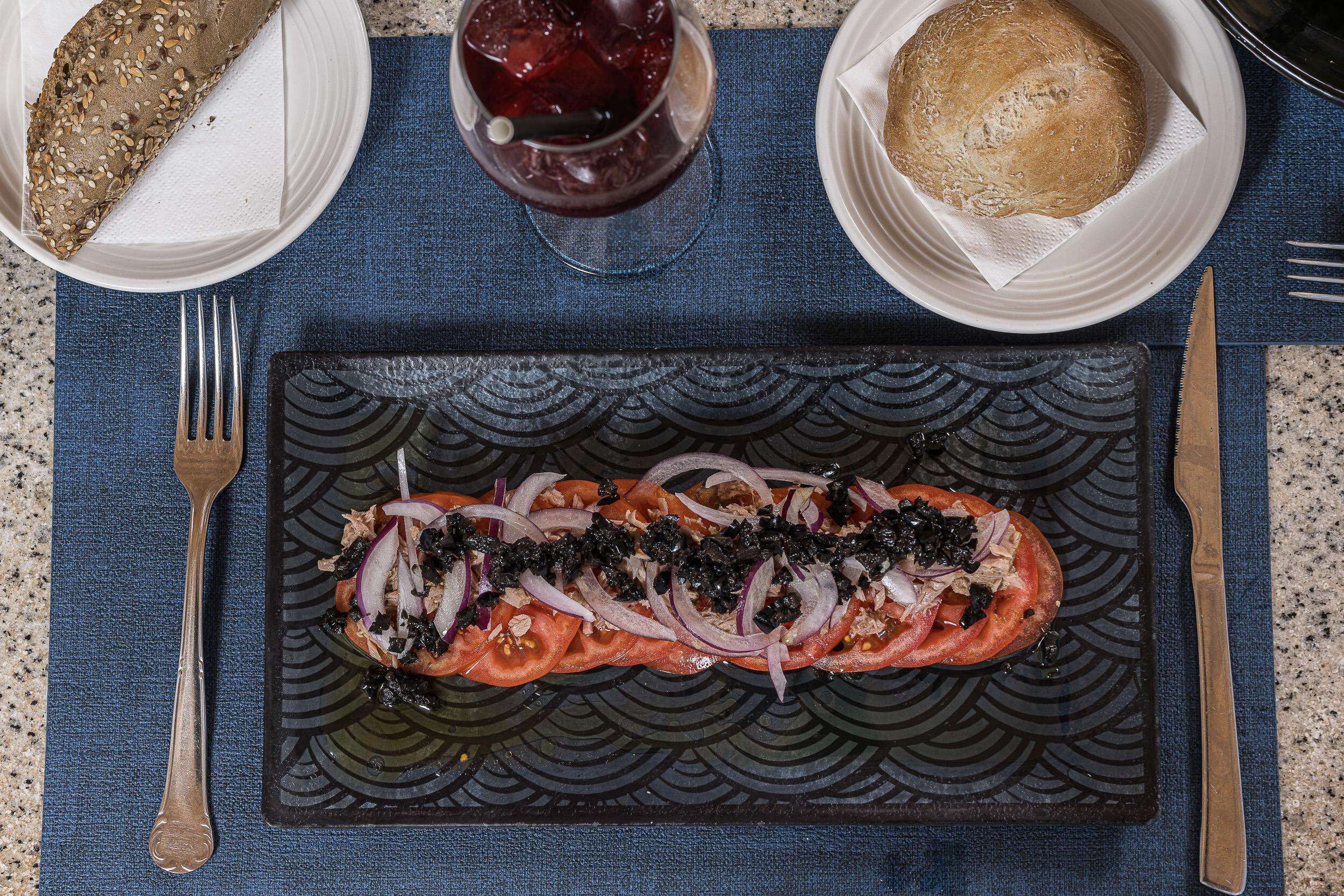 Un plat composé d’un filet de saumon, de tomates, d’oignons et de caviar noir, servi avec du pain et un verre d’une boisson rouge, le tout posé sur une nappe bleue.