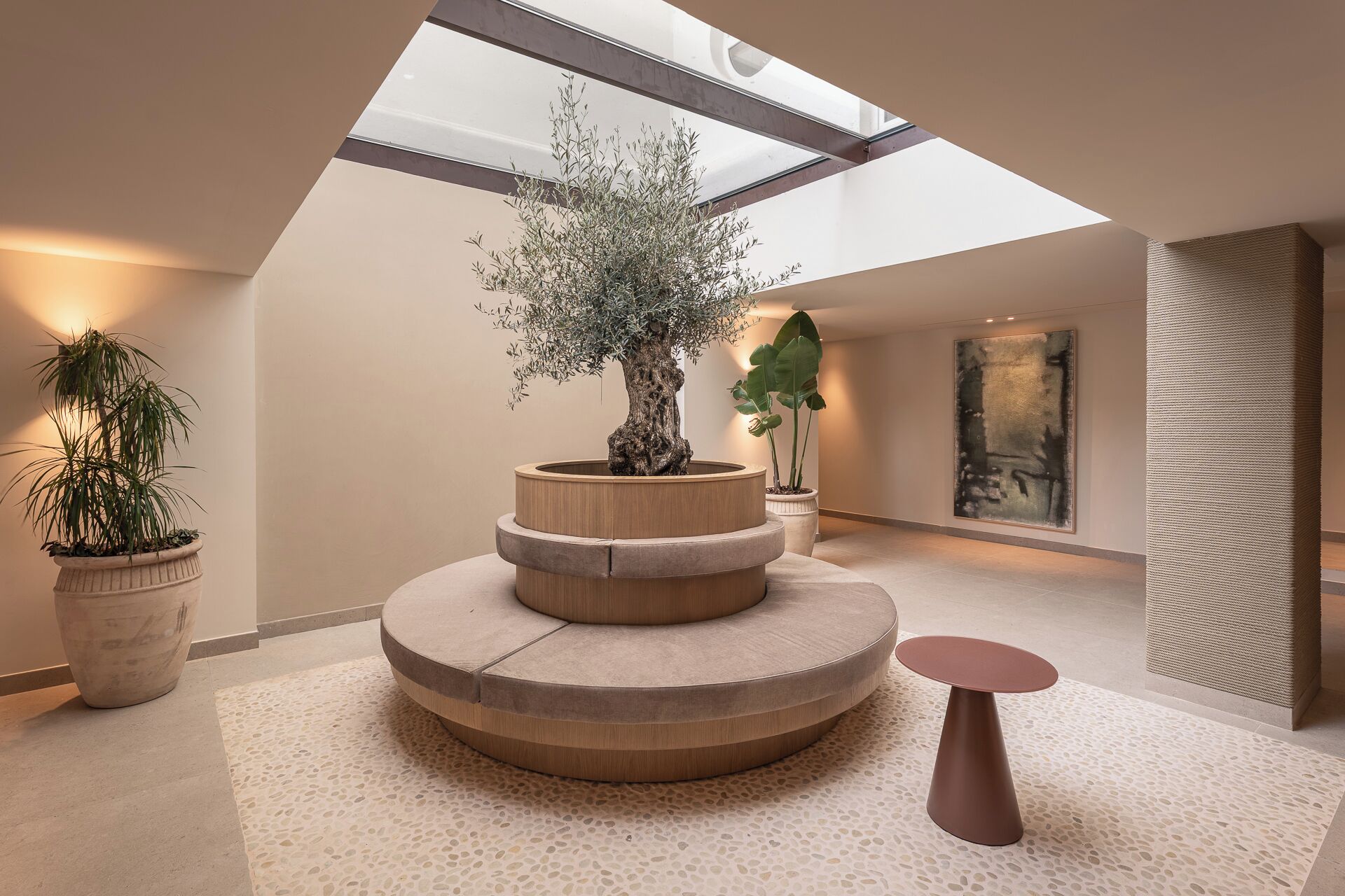 A modern interior with a skylight, a tiered wooden planter with plants, a round table, and a large abstract painting.