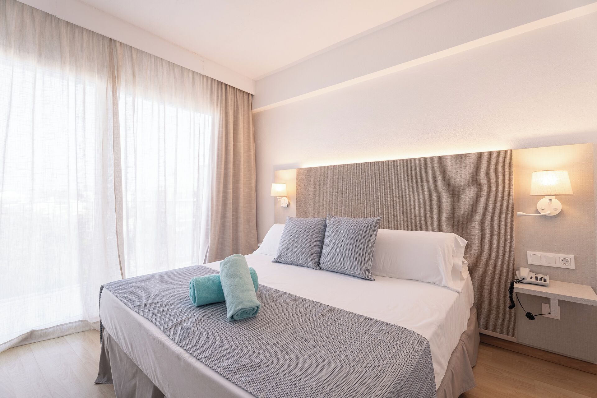 A hotel room with a bed, curtains, a headboard, wall-mounted lights, and a bedside table with a lamp.