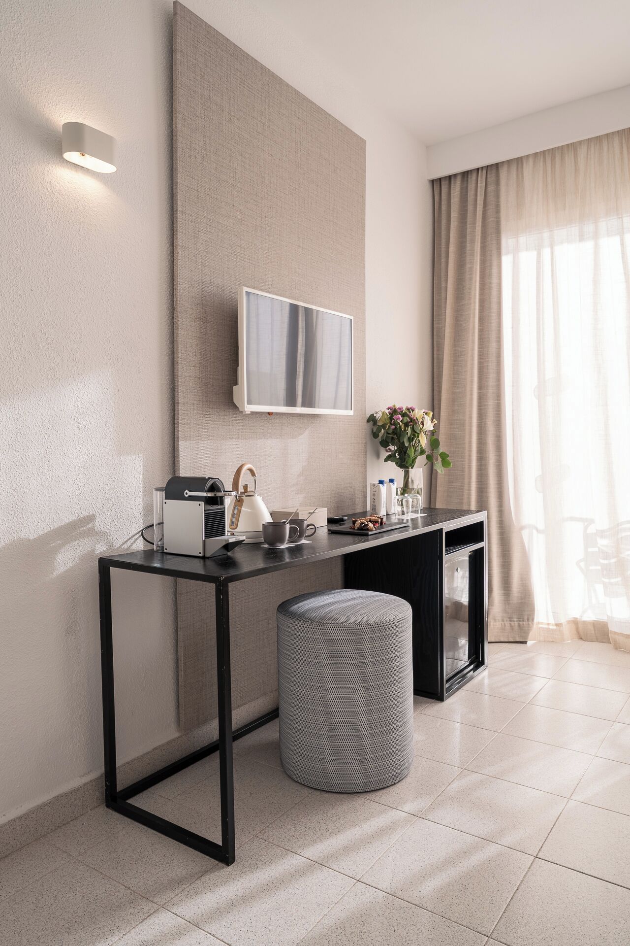 Une chambre d’hôtel moderne dotée d’un téléviseur à écran plat fixé au mur, d’une petite table avec une bouilloire et des tasses, d’une poubelle et d’une grande fenêtre avec des rideaux.
