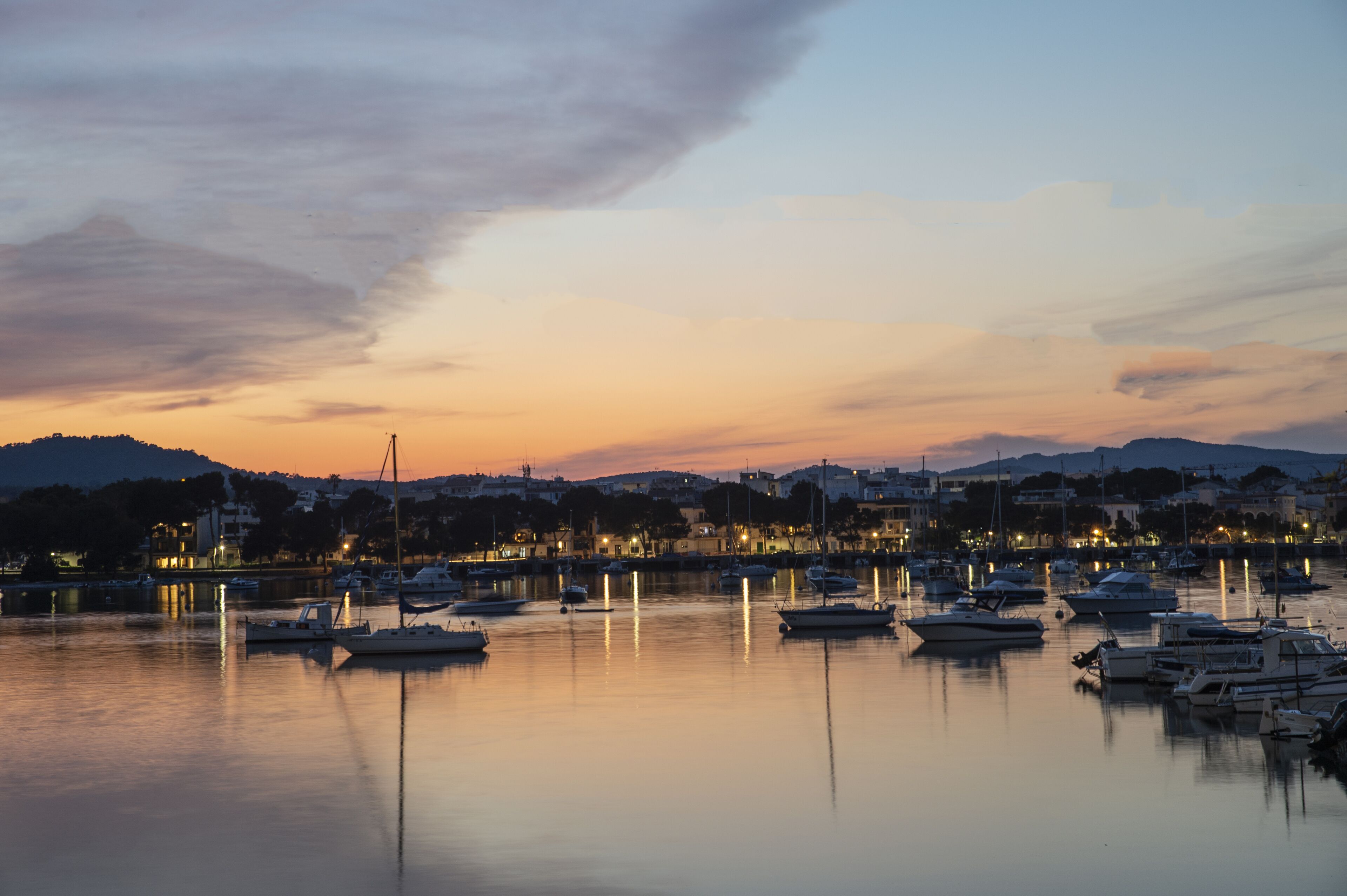 A marina at dusk with boats docked, calm water, and a cityscape in the background.