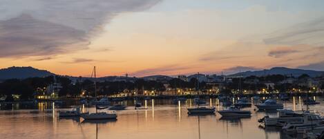 A marina at dusk with boats docked, calm water, and a cityscape in the background.