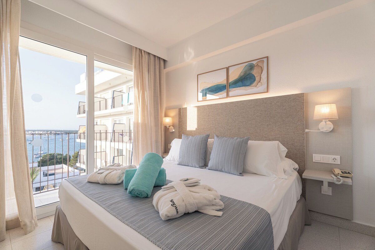 Une chambre d’hôtel avec un lit, deux oreillers, une table de chevet avec une lampe, un téléphone et une vue sur la mer.
