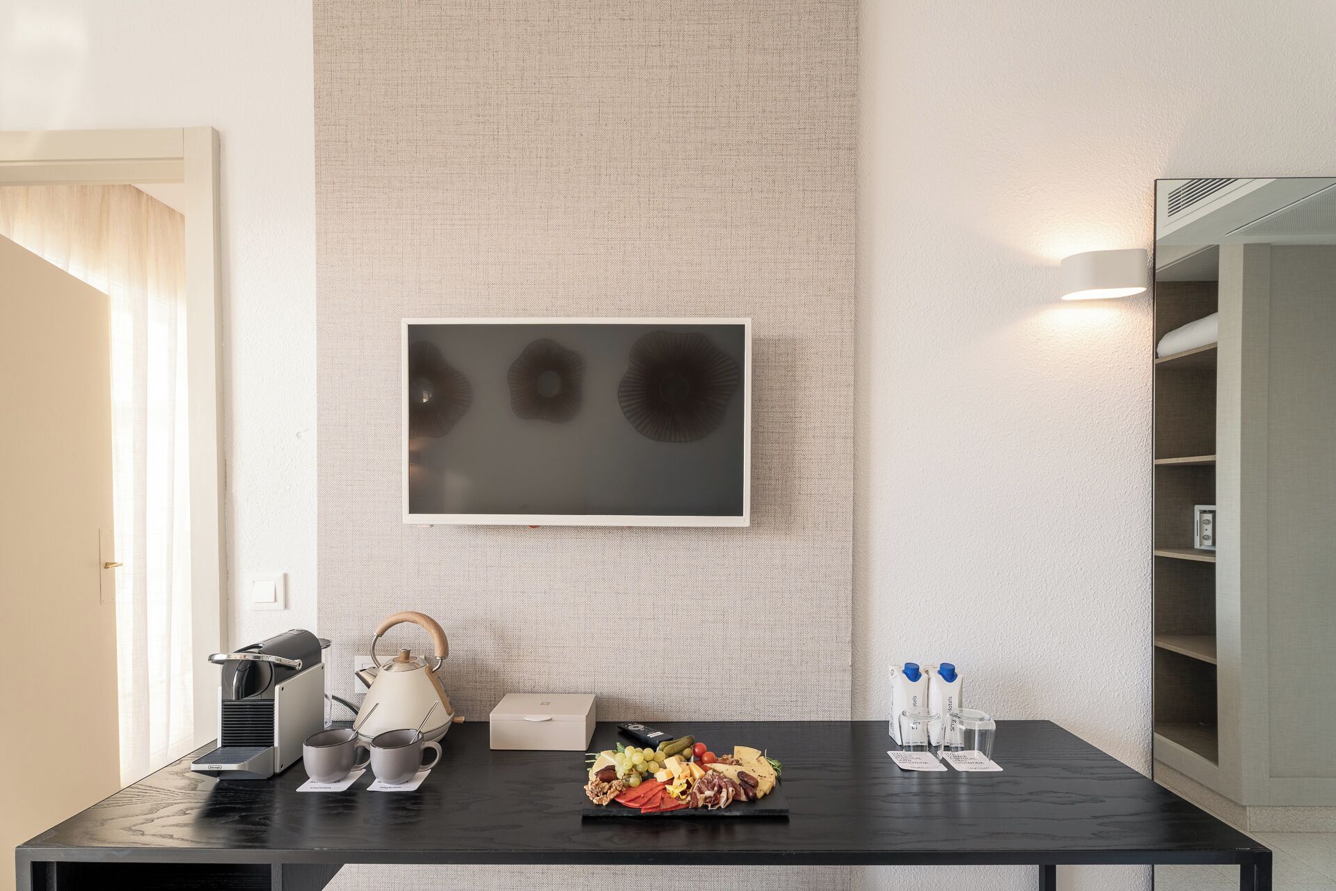 A modern hotel room with a black table, a tea set, a framed picture, and a buffet with cheese, grapes, and other items.
