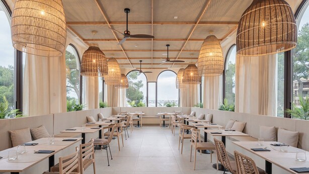 Un restaurant doté de nombreuses tables et chaises, de grandes fenêtres et de lampes en osier suspendues au plafond.