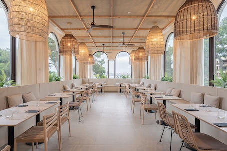 A restaurant with multiple tables and chairs, large windows, and hanging wicker lamps.