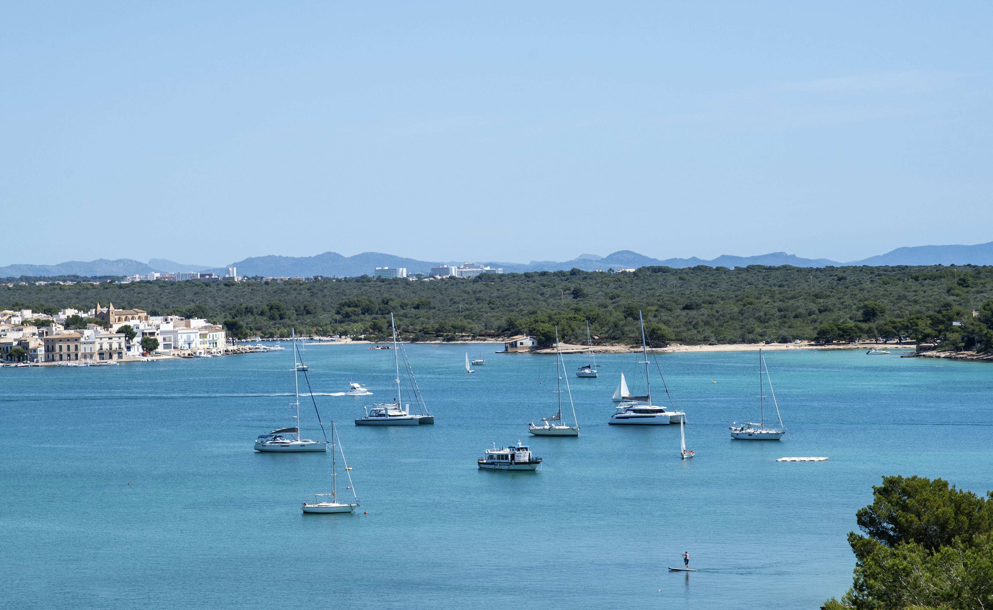 A coastal town with multiple sailboats anchored in the bay, a sandy beach, and a backdrop of hills and buildings.