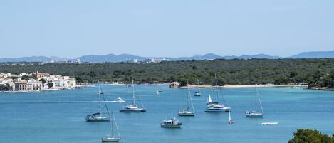 A coastal town with multiple sailboats anchored in the bay, a sandy beach, and a backdrop of hills and buildings.