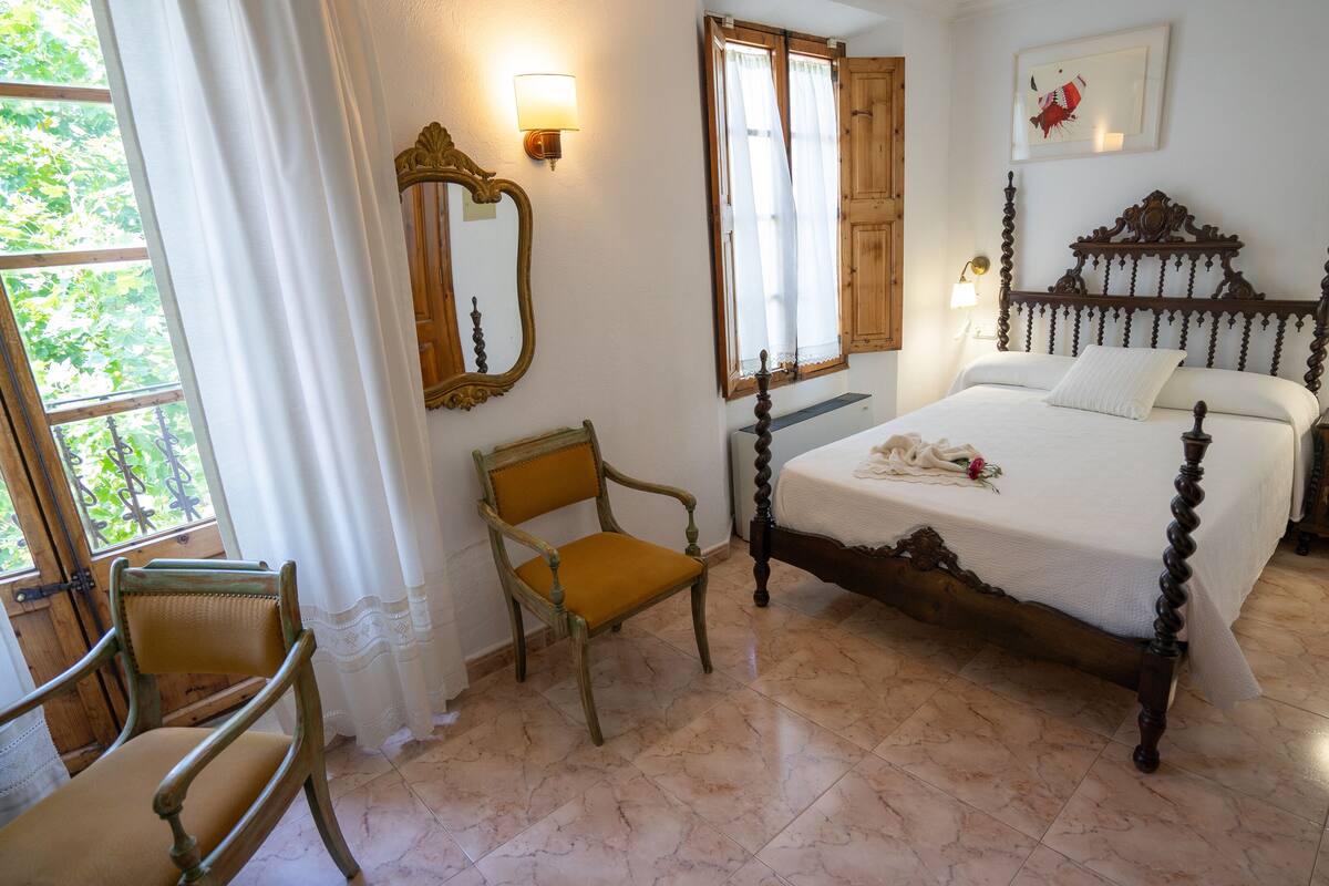 Um quarto com uma cama de dossel, uma cadeira de madeira, uma mesinha de cabeceira e uma janela grande com cortinas.