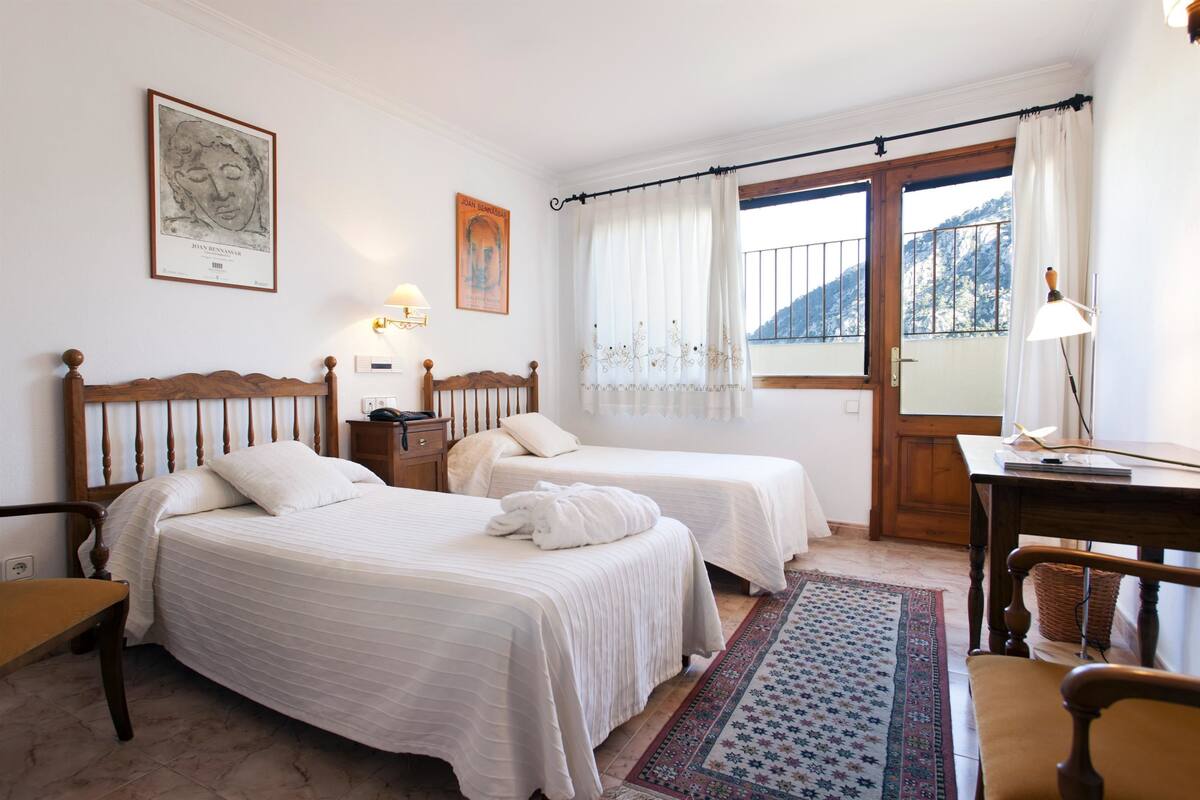 Quarto com duas camas, cabeceira de madeira, mesa de cabeceira, escrivaninha e janela com cortinas.