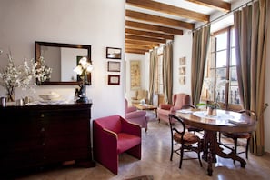 A well-lit room with a wooden ceiling, a dining table with chairs, a red armchair, a wooden cabinet, and a large window with curtains. - Hotel Juma (Pollenca)