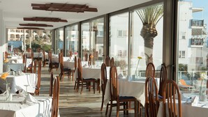 A restaurant with tables and chairs arranged for dining, large windows offering views of buildings and palm trees, and a modern ceiling design. - Hotel Abrat (Sant Antoni de Portmany)