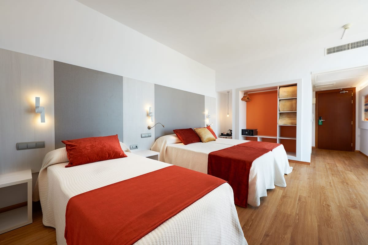 Hotellihuoneessa on kaksi sänkyä, joissa on punainen peitto ja tyynyt, valkoiset päätyosat ja puulattia. Taustalla näkyy vaatekaappi, jossa on hyllyjä ja ovi.