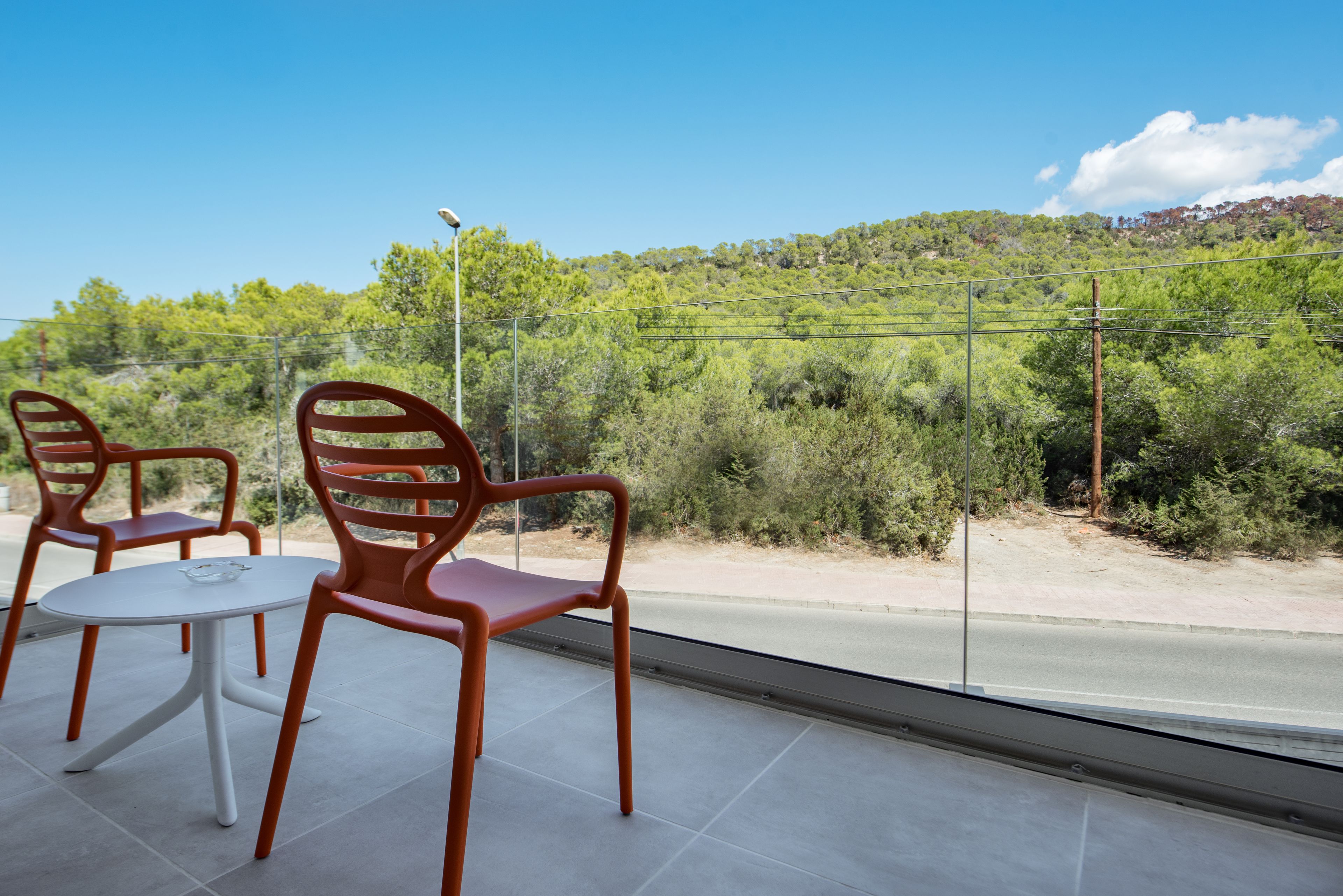 A balcony with two chairs and a table, overlooking a road and a hilly landscape with trees.