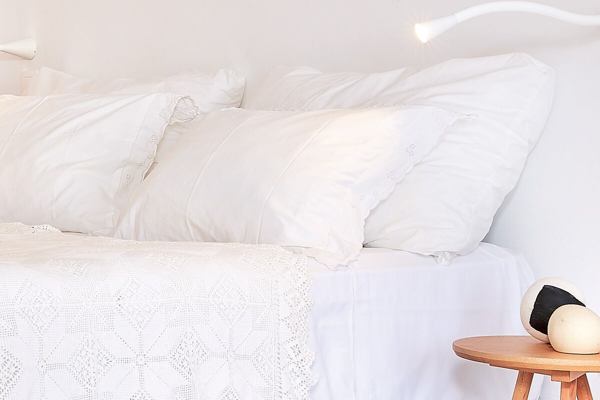 Una cama con sábanas blancas y un cubrelecho de encaje, una mesita de noche con una lámpara, y una pequeña mesa redonda con dos objetos blancos.