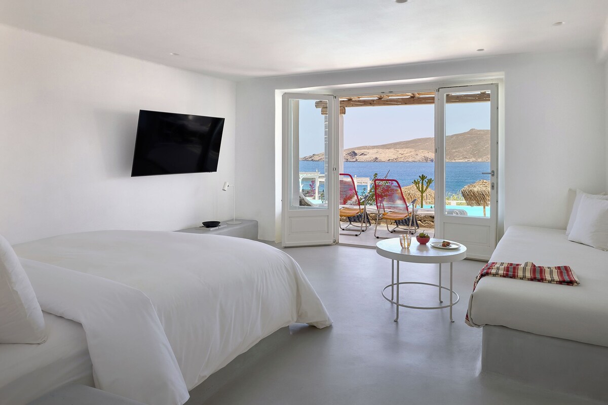 A hotel room with a bed, a TV, a small table, and a view of the sea.