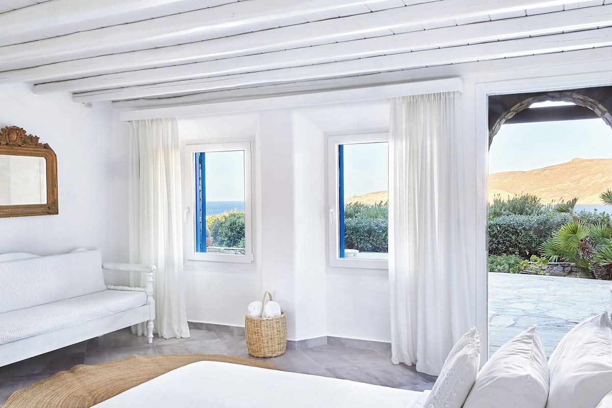 A bedroom with a large bed, two windows with curtains, a wicker basket with towels, and a view of the sea and hills.
