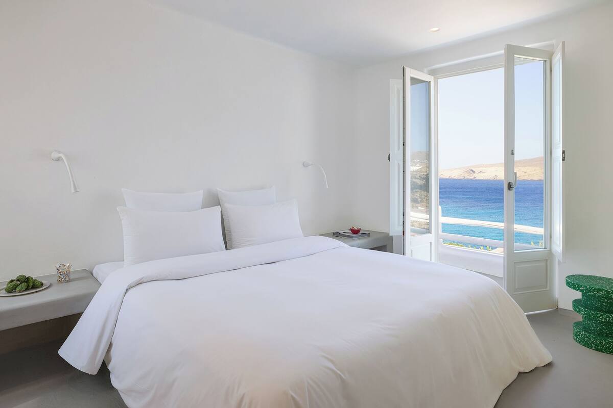 A bedroom with a bed, white walls, a nightstand, a small table with a book and a red bowl, and a green stool.